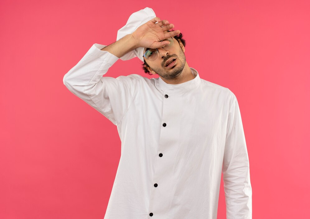 Exhausted chef in white uniform wipes brow, illustrating how ghost kitchen work can be demanding, on pink background.